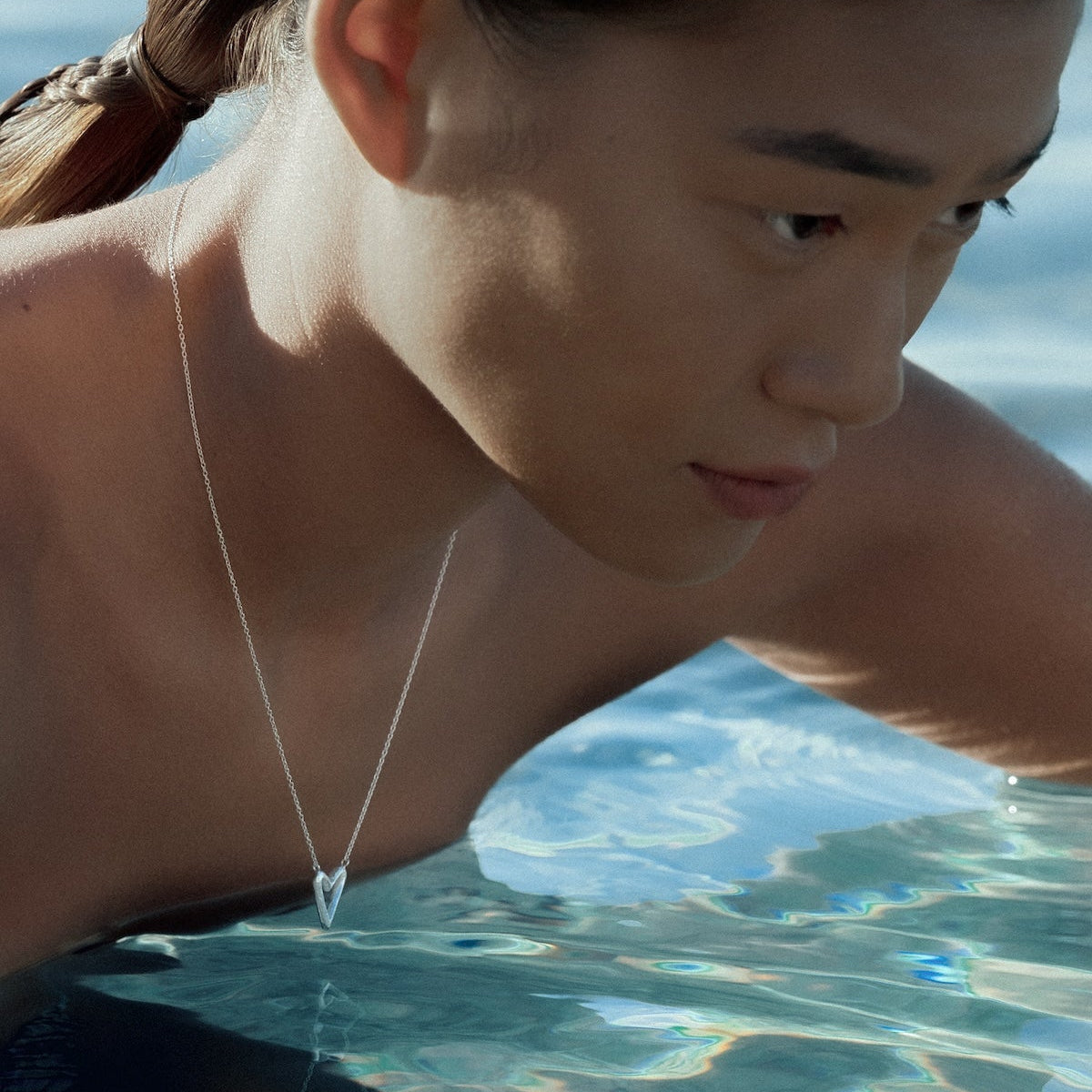 A close-up lifestyle shot of a model in shallow water wearing a delicate silver heart pendant necklace.