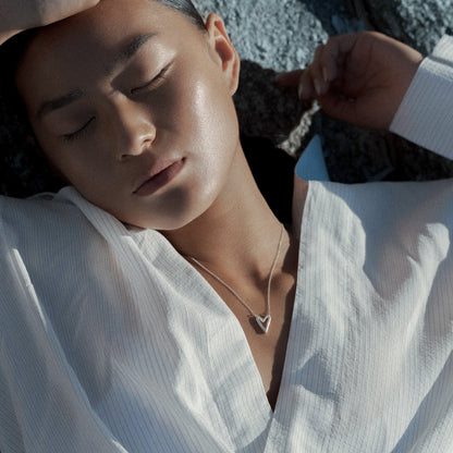A close-up lifestyle shot of a person lying on a dark textured surface, wearing a white button-up shirt and a silver necklace with a heart-shaped pendant.