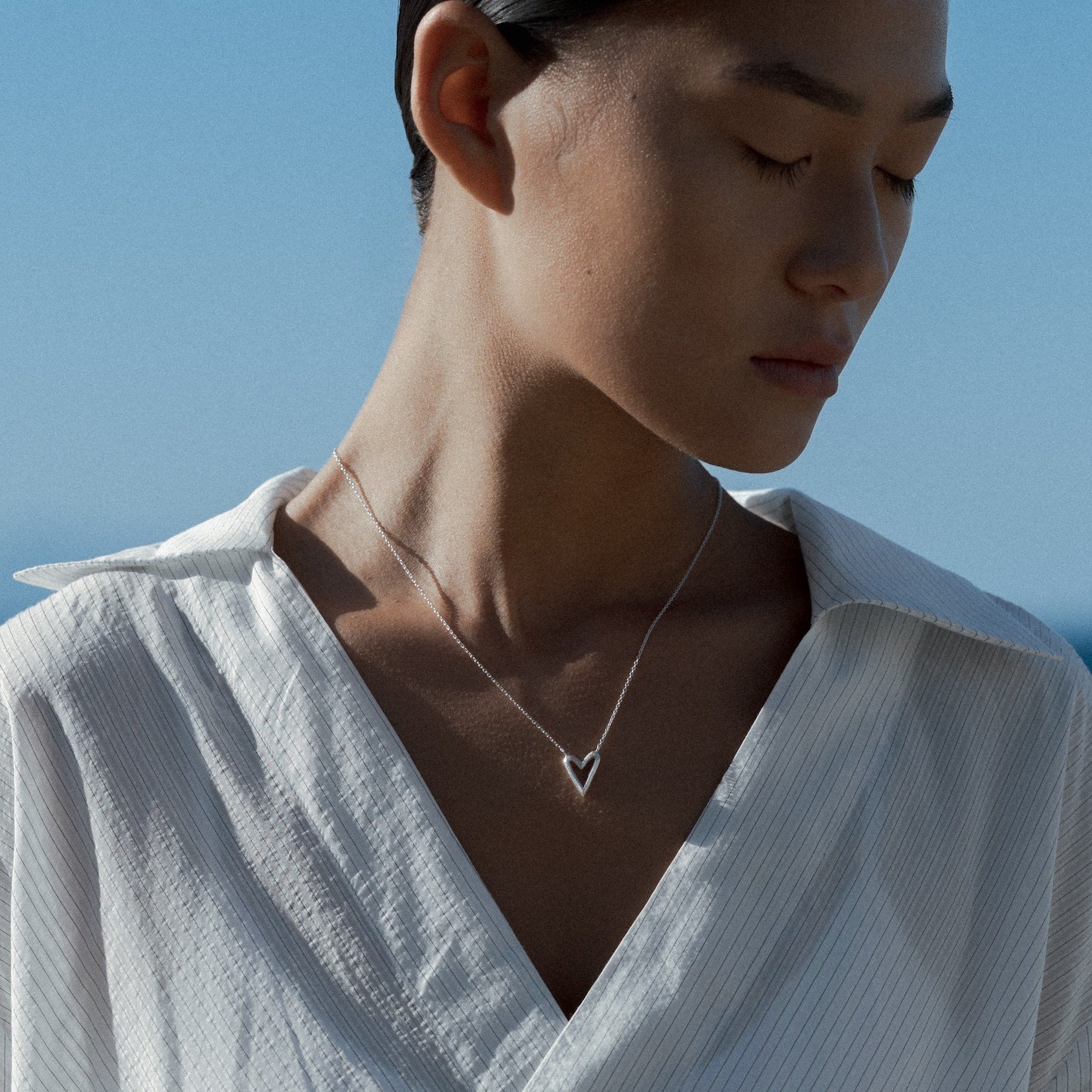 Person wearing a white shirt with a small silver heart necklace, standing against a blue sky and ocean.
