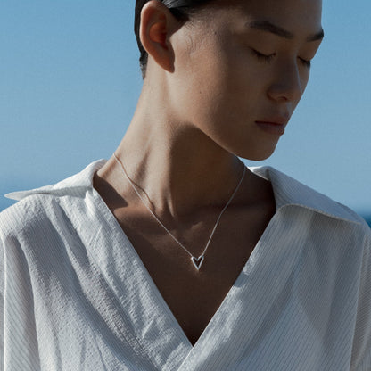 Person wearing a white shirt with a small silver heart necklace, standing against a blue sky and ocean.