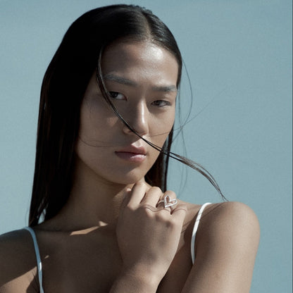 A woman with long dark hair wearing a white spaghetti-strap camisole top and a silver heart ring, posing with her arms crossed against a light blue background.