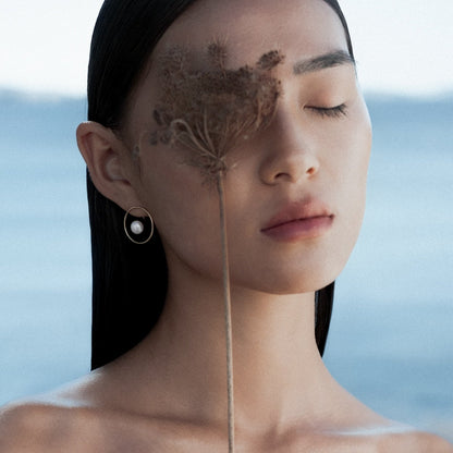 Woman with a flower on her face wearing a Calliope pearl earring by Aurore Havenne against a blurred natural background of the sea in Mykonos