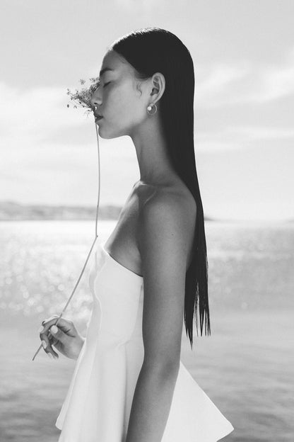 A black and white portrait of a woman in profile wearing a strapless white dress, holding a delicate flower stem near her face and wearing the Pearl Calliope earrings by Aurore Havenne, with a soft blurred outdoor background.