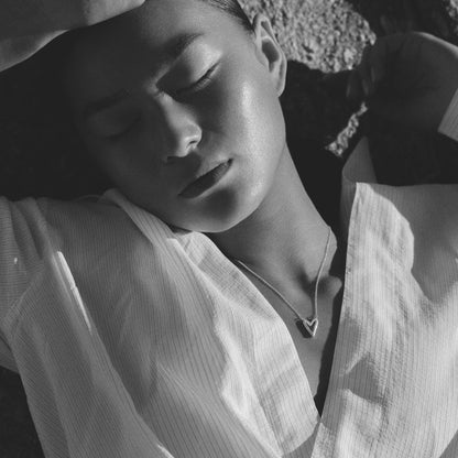 A black and white close-up photo of a person lying down with arms behind their head, wearing a light-colored, open shirt and a heart necklace.