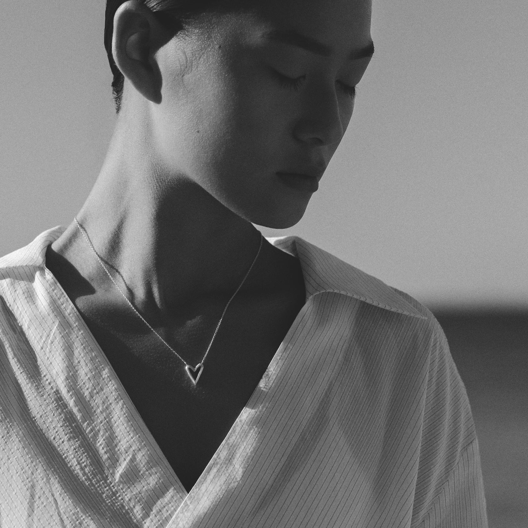 A black and white close-up portrait of a person wearing a white textured V-neck shirt and a delicate necklace with a small heart pendant, set against a soft, blurred background.