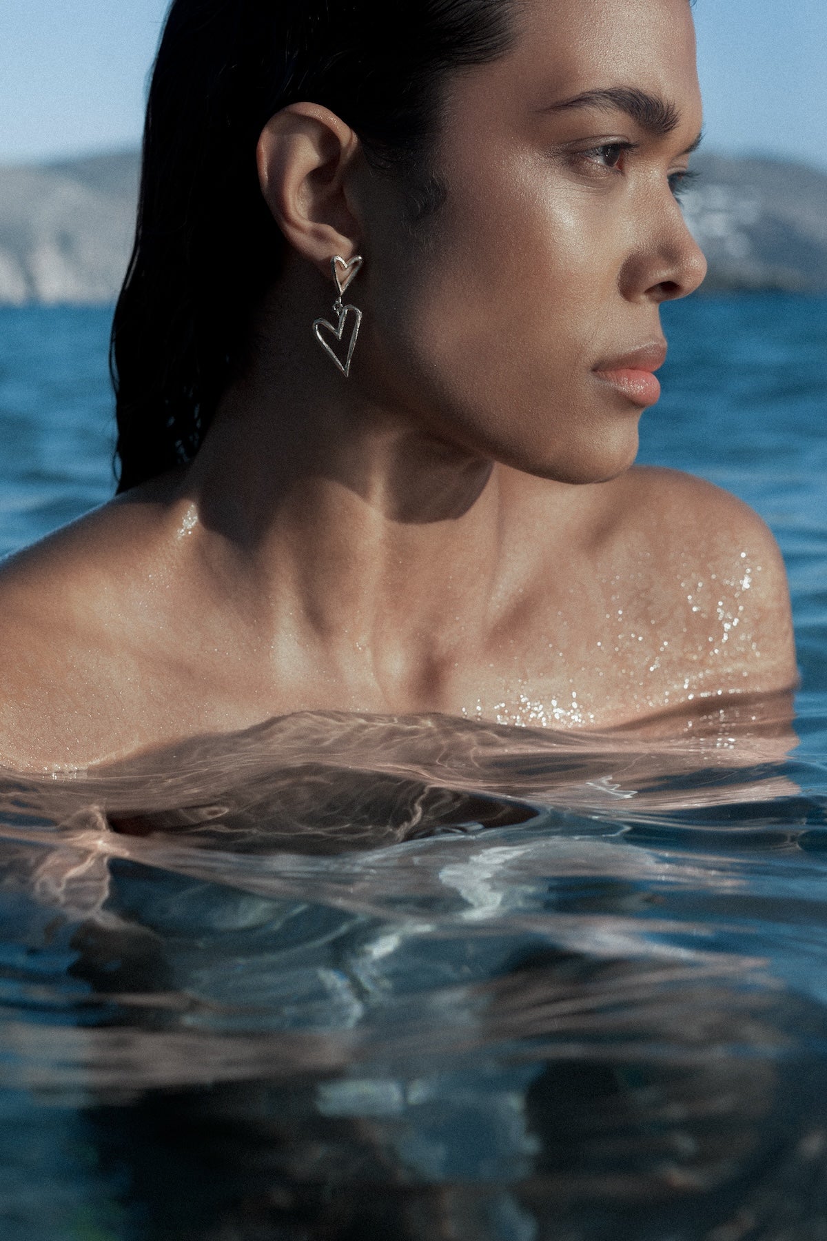 A woman Livia Eduarda in shallow water wearing a dangling heart-shaped earring, with blue water and a distant shoreline in the background.