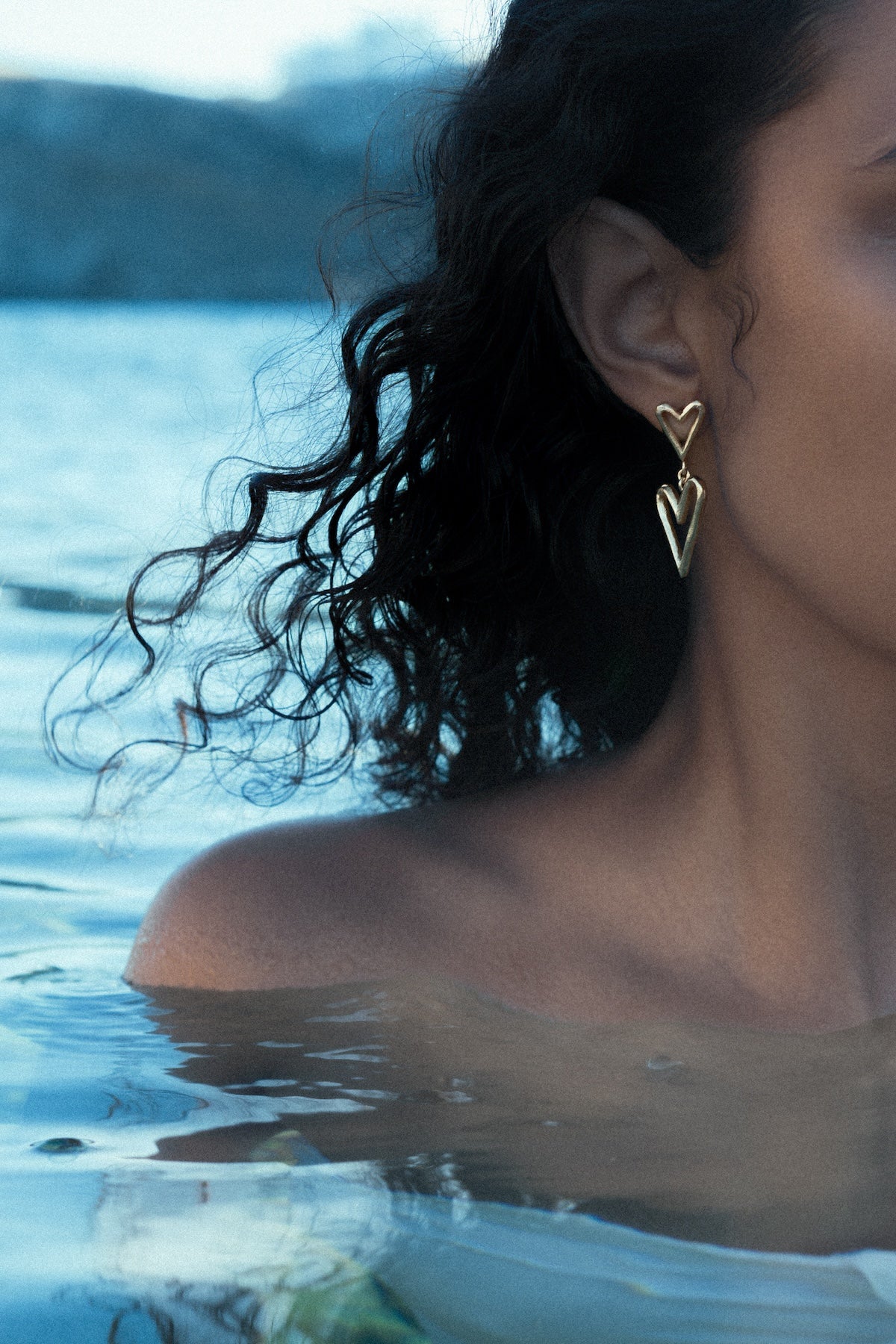A close-up lifestyle shot of a woman with curly dark hair wearing a gold heart dangle earring in the sea in Mykonos